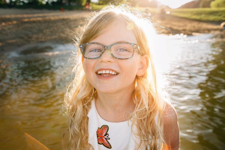 Young girl with glasses and curly blonde hair standing in the water at a park beach with the sun setting over her head.