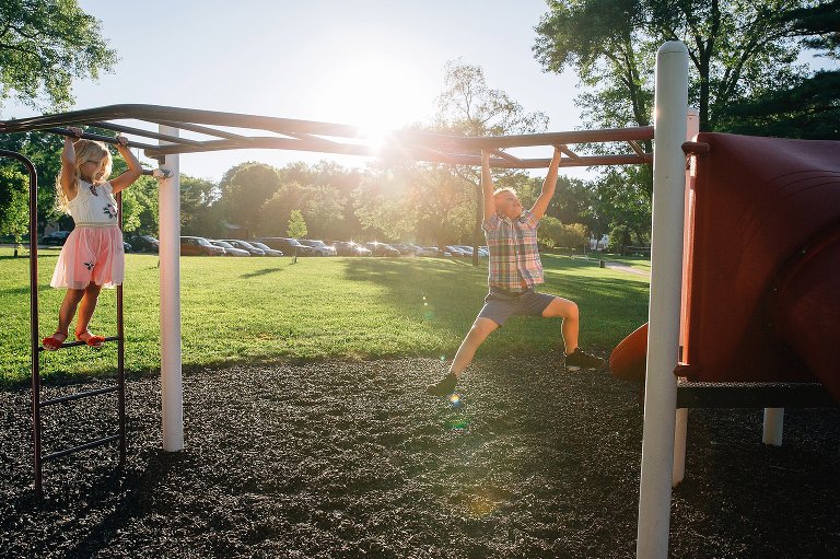 Young boy climbs across the monkey bars at a park with his little sister waiting after him.