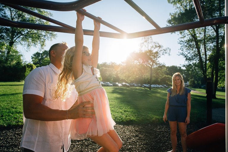 Father holds up daughter helping her across the monkey bars at a park, mom standing and watching from behind.