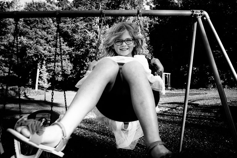 Black and white. Young girl with glasses and long curly hair swings on a swing set at a park.