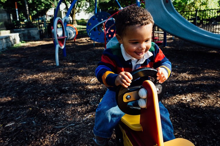 Young toddler boy rides a playground motorcycle. He's smiling and slightly sticking out his tongue. 