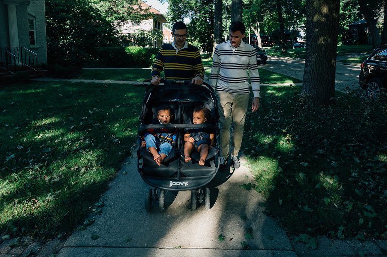 two men walk their two kids in a stroller through a residential neighborhood. 