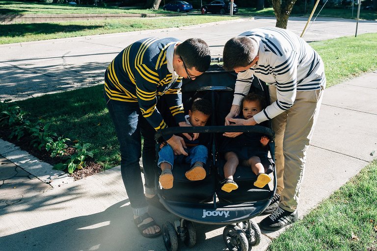 Two dad bend over to buckle in their two kids in a double stroller. 
