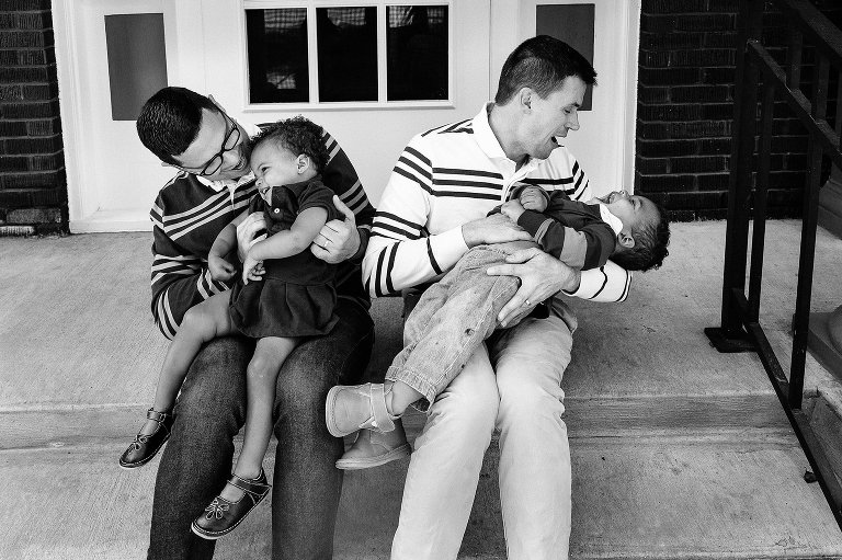 Black and white. Two dads hold and laugh with their two kids. 