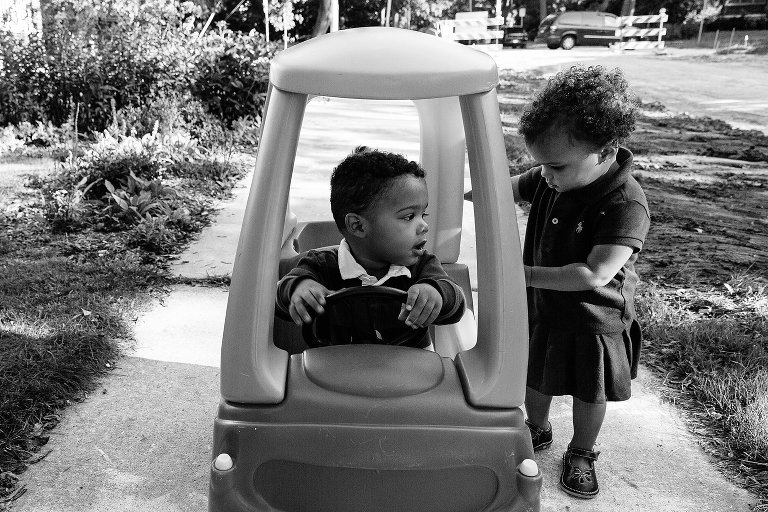 Black and white. Toddler girl tries to open the door to a toy car that her brother is riding in.