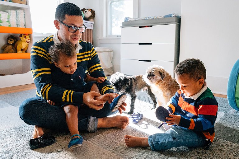 Dad and two young kids and two dogs sit on the bedroom floor putting on shoes. 