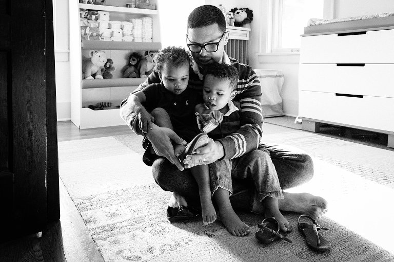 Dad helps daughter put shoes on her feet. His daughter and his son are both sitting on his lap.