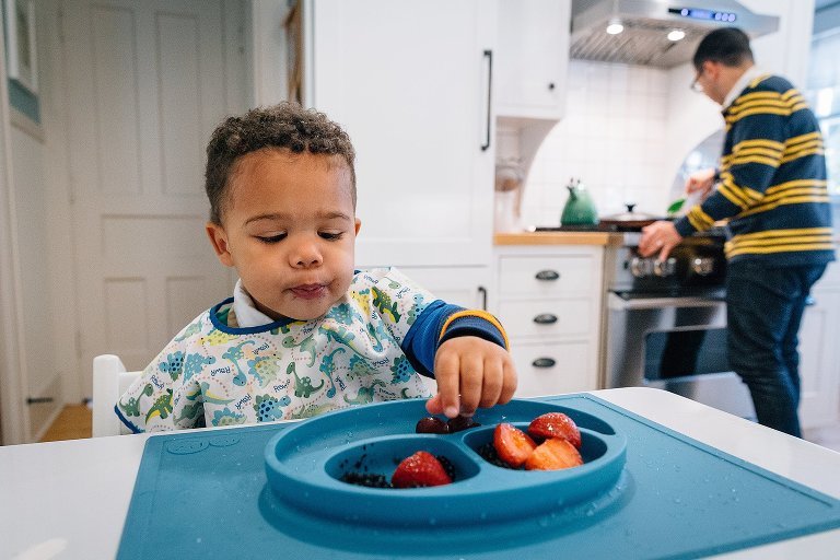 Toddler boy reaches for a grape on his plate while Dad works at the stove behind him. 