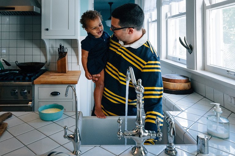 Toddler girl laughs as dad runs the kitchen sink faucet. 
