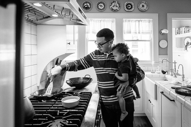 Black and white. Dad holds girl on his hip while using spatula in a pan with other hand. 