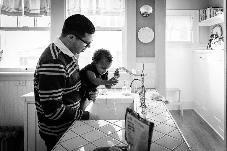 Black and white. Dad holds daughter while she washes her hands in the kitchen sink. 