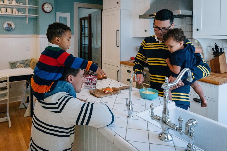 One dad lets son sit on his shoulders while they watch Dad #2 prepare eggs with toddler girl. 