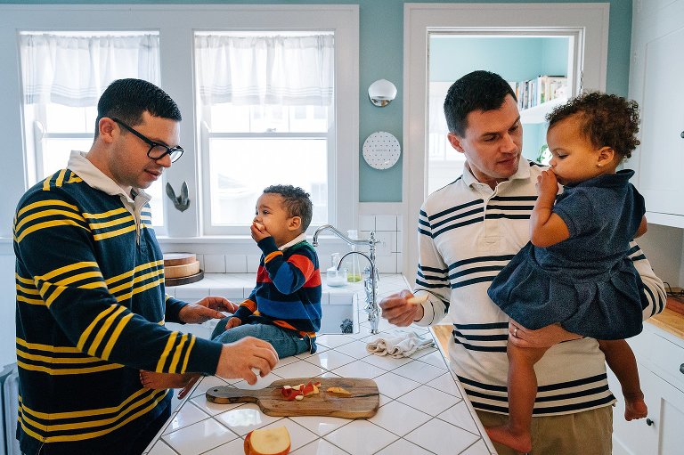 Two dads help prepare an apple snack for their two toddler kids. 