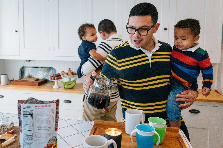 One dad pours coffee in the bright white kitchen and holds son, second dad holds daughter in the background while getting a snack. 