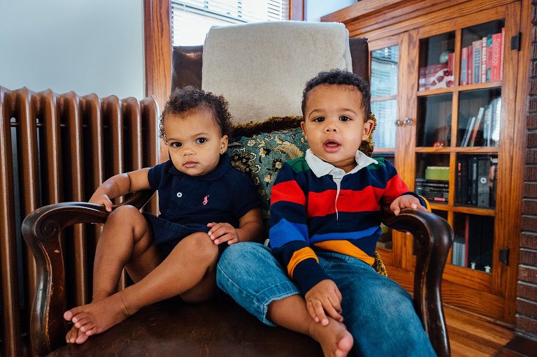 A boy and girl toddler sit on chair together. They are camera aware. 