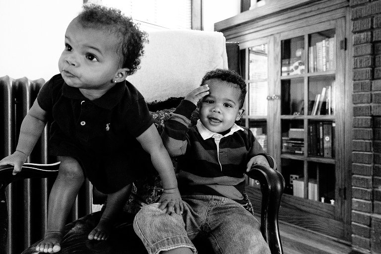 Black and white. Young toddler girl uses her brother to stand up. Brother touches his head from a bump and smiles. 