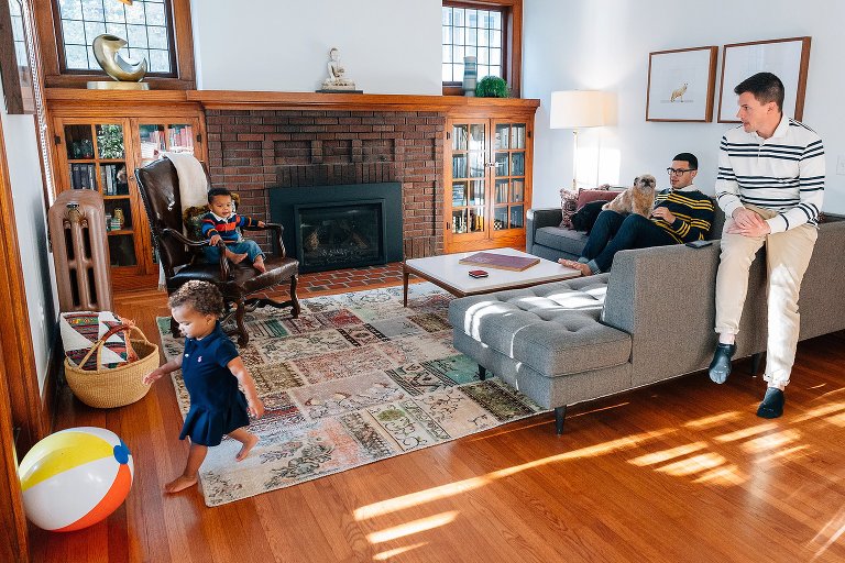 Two dads and their two kids and a dog play in a bright, cheery living room together. There is a large beach ball in the space. 
