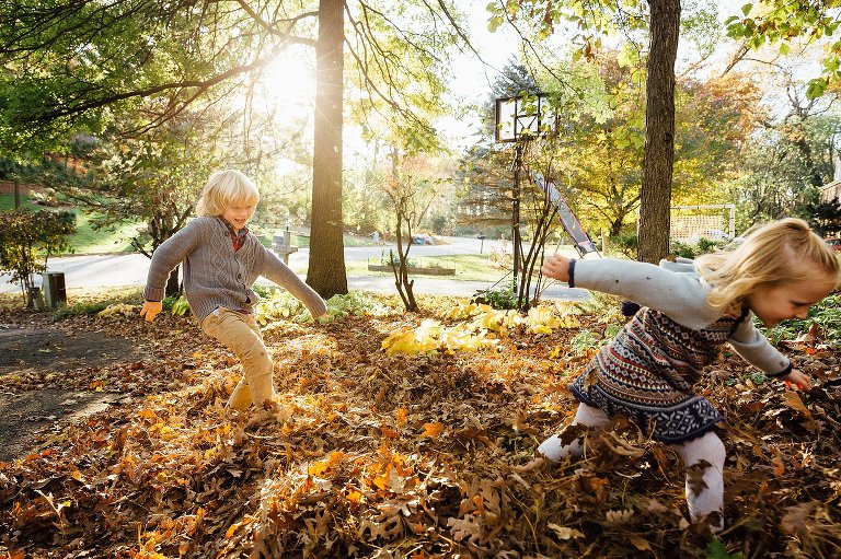 boy kicks leaves toward his sister running away during Autumn at Home photo session