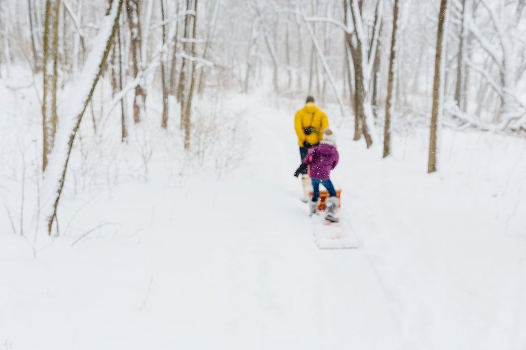 Purposeful out of focus shot of snow falling in the woods with dad and daughter pulling/riding a red sled in the distance. madison family photographer 
