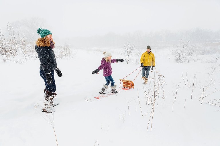 Girl rides sled with outstretched arms as if riding a surf board. Her father is pulling the sled and her mother is behind them. madison family photographer 