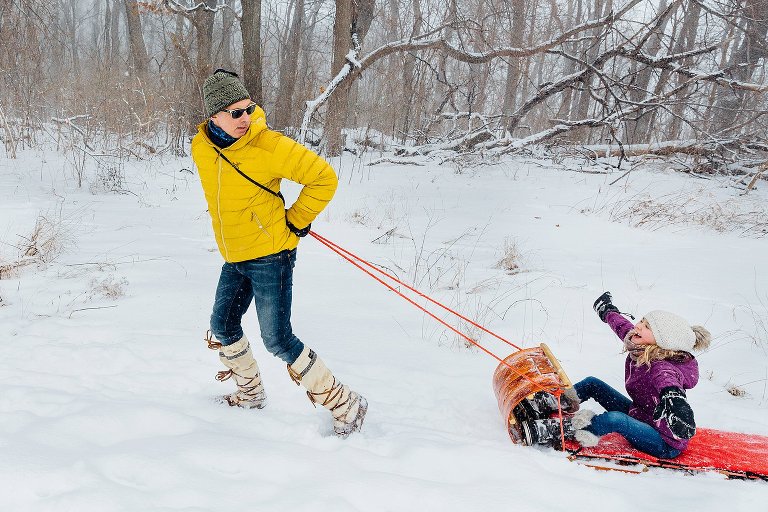 Girl holds arms out while riding on a sled. Dad pulling sled and looks behind at daughter. madison family photographer 