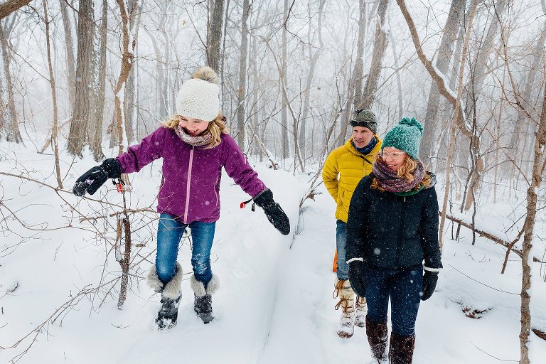 Girl walks on a fallen log--arms out for balance--while her parents walk beside her. They are in a snowy forest. 
