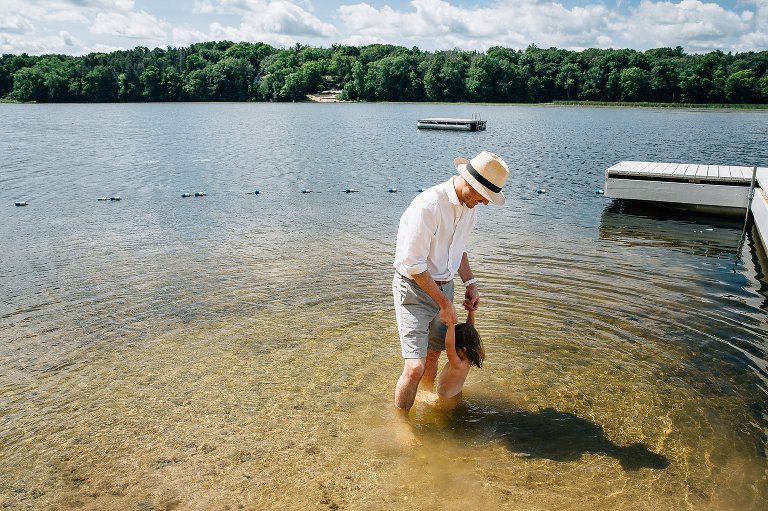 Father holds daughter's hands while she takes a dip in the lake. Big puffy white clouds and green trees in the distance. 