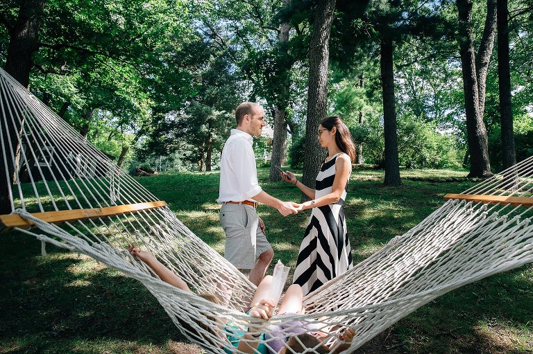 Mom holds phone and looks at screen while holding dad's hands and girls in hammock in foreground. 