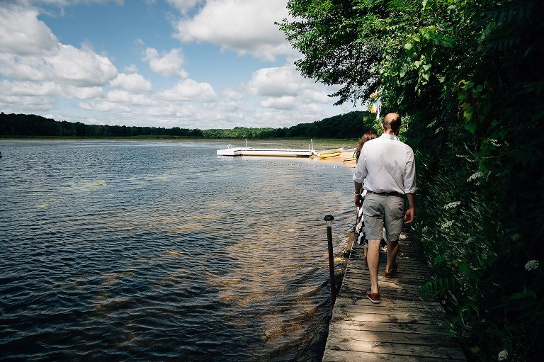 Family walks on narrow boardwalk between lake and land. White clouds and bright blue sky is reflected in water ripples. White pier is in the distance.