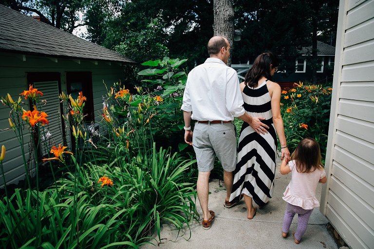 Dad reached out to mom while mom holds daughter's hand as they walk among orange day lilies. 