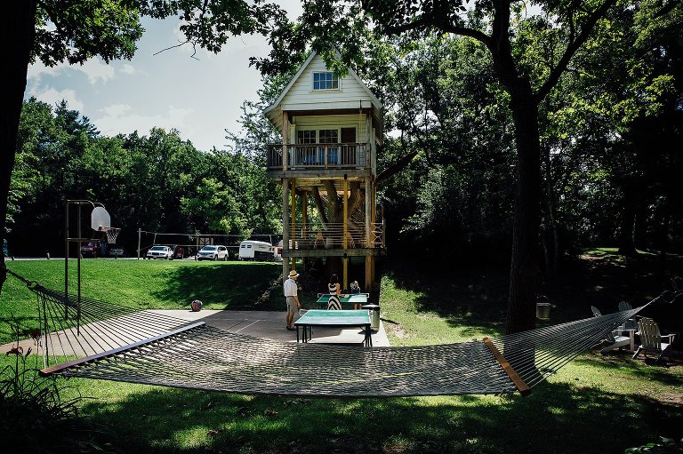 Mom, dad, two daughters play ping pong with hammock in foreground and treehouse in background of summer campground. 