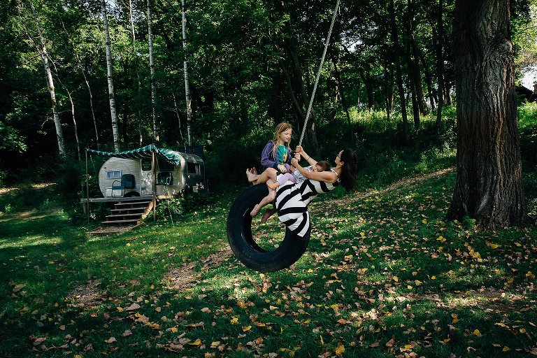 Mom, two girls ride on tire swing. Small camper in the background. 