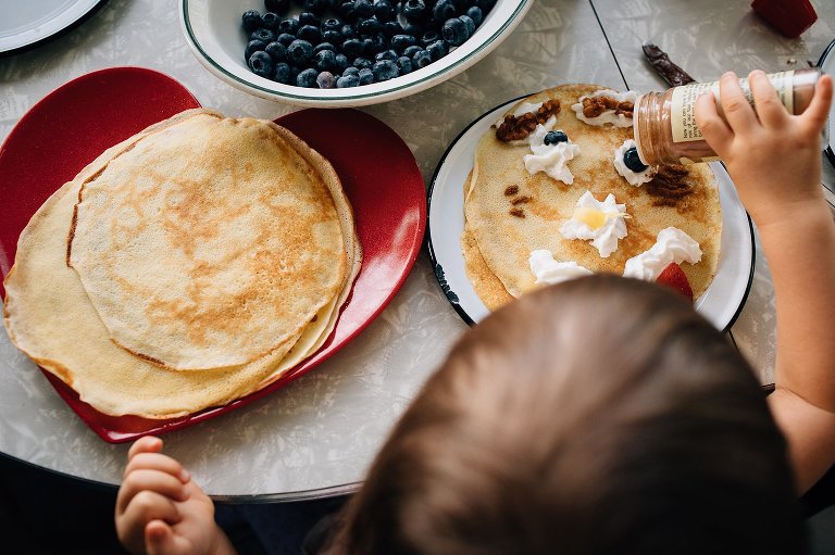 Small child sprinkles cinnamon on crepes decorated with a whipped cream and fruit "face" 