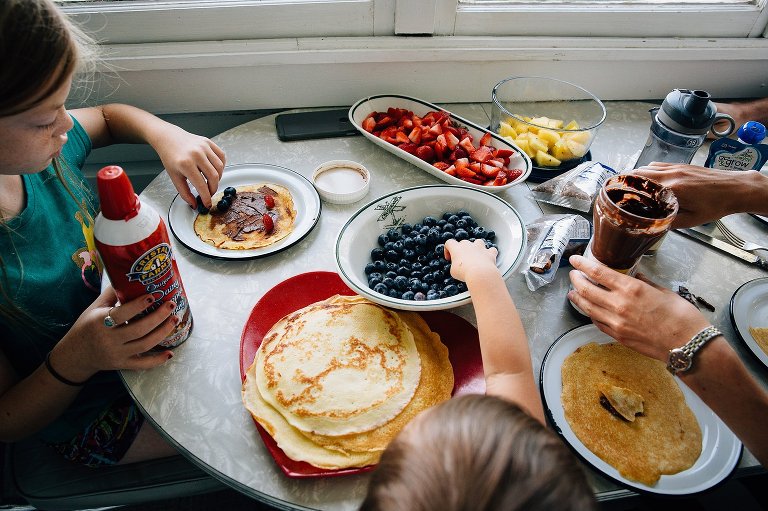 Vibrant breakfast table scene: blueberries, strawberries, crepes, whipped cream, Nutella and hands all reaching for food items. 