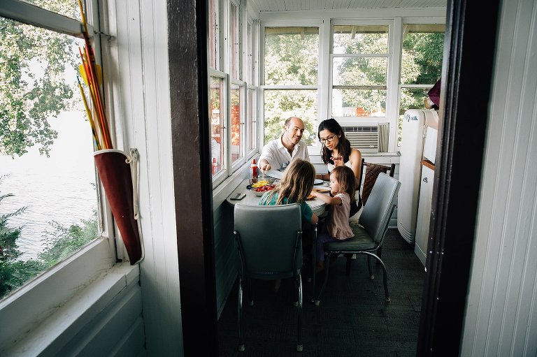 Family of 4 eats breakfast in old-fashioned summer cabin next to trees and lake. 