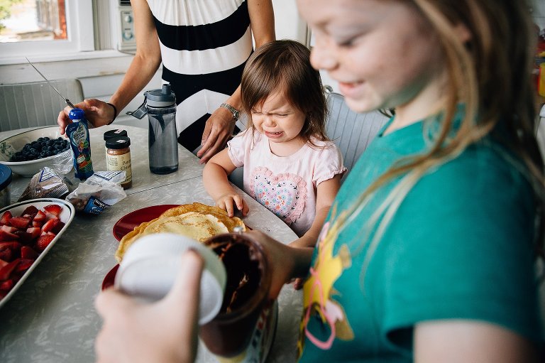 Family at breakfast table. Young daughter cries as older sister smiles opening a jar of Nutella.