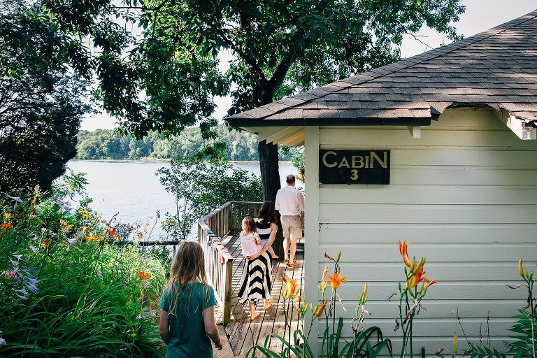 Family of 4 walks back to cabin overlooking a lake. Summer flowers everywhere. Mom holds 2yo on her back. 