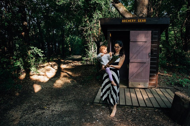 Mom and young daughter come out of archery hut in the woods. 2yo holding an arrow.