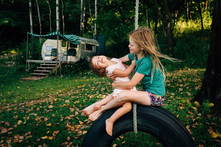 Older sister holds younger sister on tire swing. Wind blows their hair.