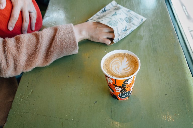 Fancy latte in foreground, hand reaching into bag with cookie in background. 
