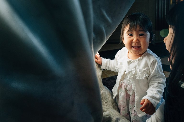 1yo girl stands next to statue with sister. she makes eye contact and smiles with camera.