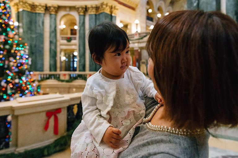 One year old girl looks at her mother and hols her necklace. Large Christmas tree in the background .
