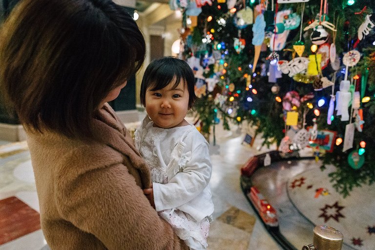 Mother holds one year old girl in front of large Christmas tree