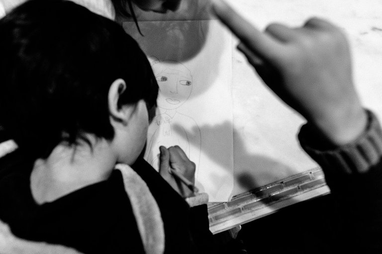 black and white. older girl draws while younger child points above her, casting a finger pointing shadow on her paper. 