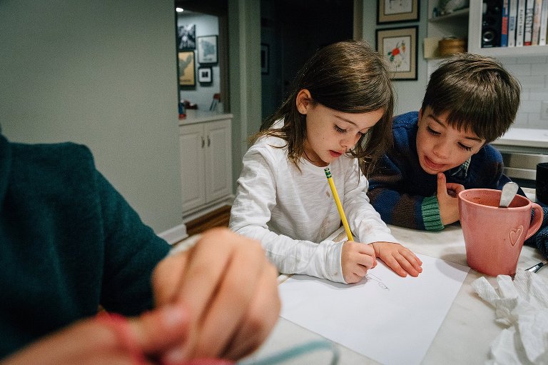 one 5yo draws while 5yo twin watches. Older sister does craft with her hands in the foreground. Kids siting at kitchen island. 