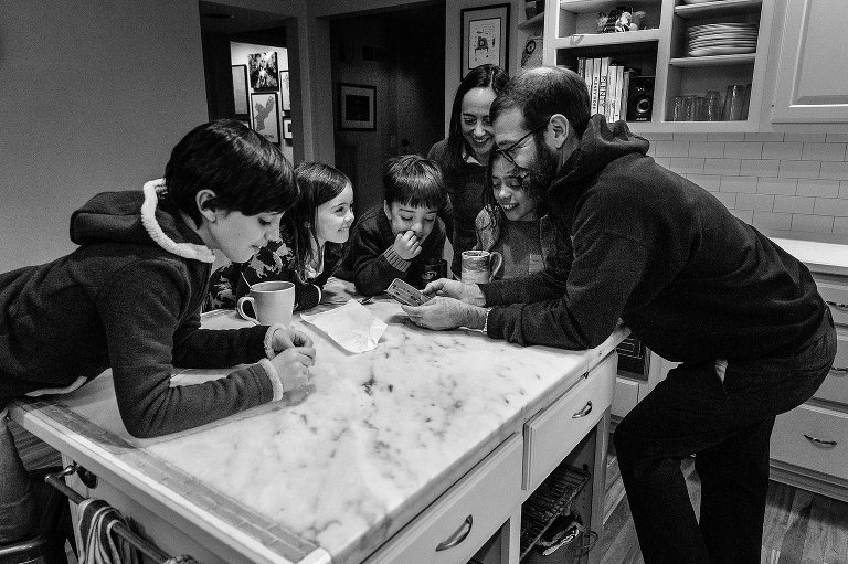 Family of 6 gathers around kitchen island to share old videos and laugh. 