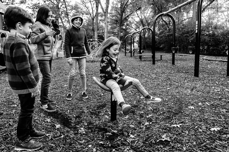 Black and white. 5yo spins on playground equipment while 3 siblings cheer and watch. 