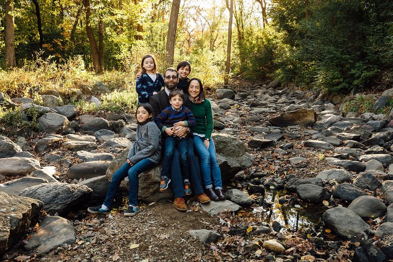 Family of 6 sits in and stands in a dry creek bed for a family portrait. Fall leaves and late sun in the background. 