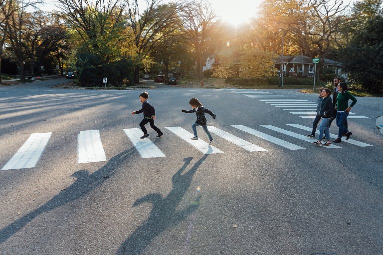 Two young kids skip across crosswalk, long shadows across the image. the rest of the family of 4 follows in a cluster behind them.