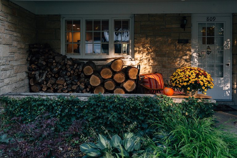home with stacked firewood, pumpkins, and large yellow mums on porch. Dappled sunlight hits the house. 